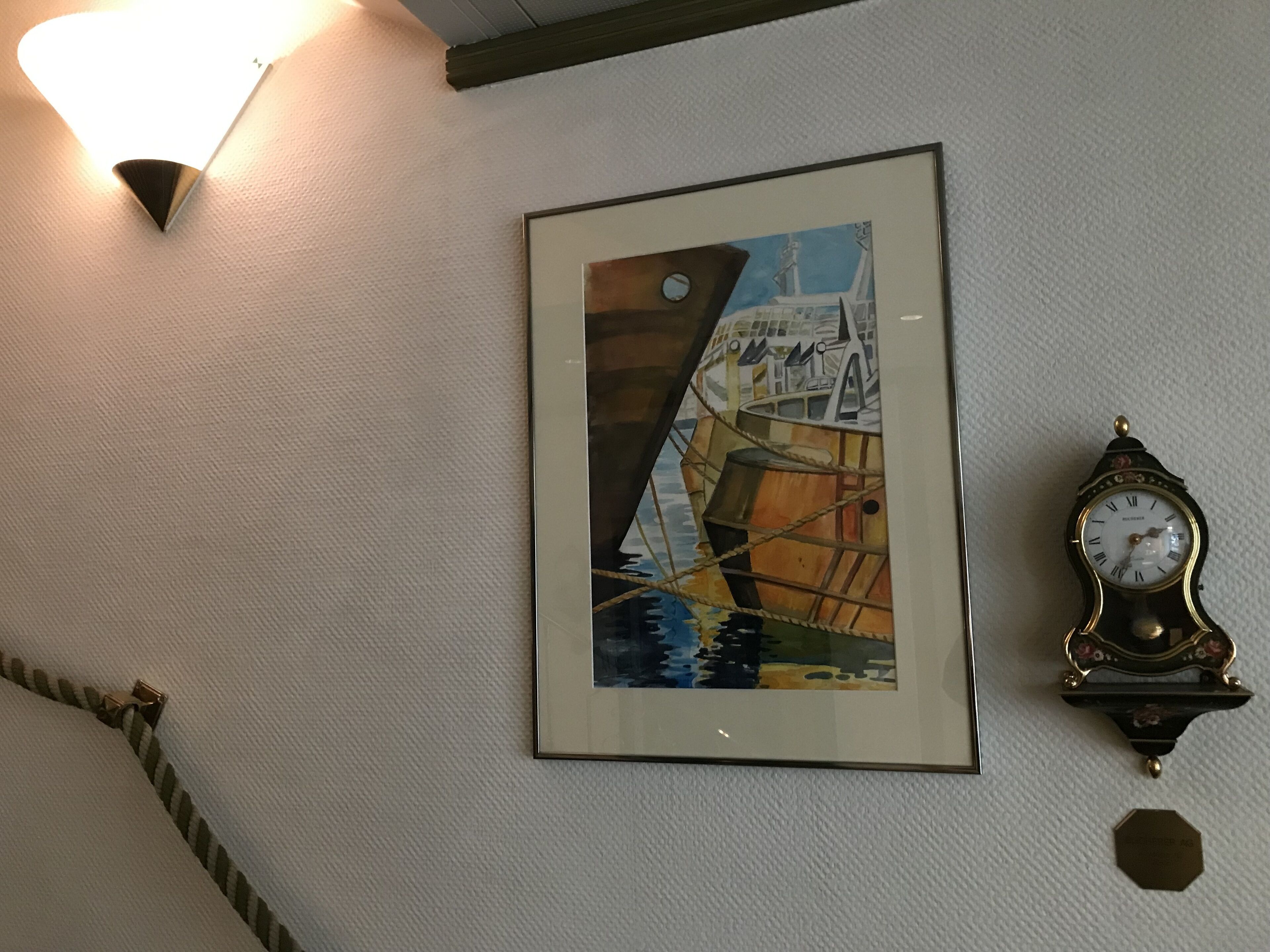 Un cuadro enmarco con un barco en la pared, un reloj sujeto a la pared y una lámpara.