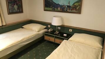 A hotel room with two single beds, a nightstand with a lamp, a phone, and a painting of a mountain village on the wall.