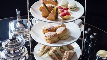 Un soporte de tres niveles para postres con varios pasteles y sándwiches, acompañado de té y condimentos sobre una mesa reflectante.