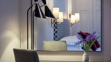 A hotel room with a desk, chair, and a large mirror reflecting a bed and a vase with flowers.