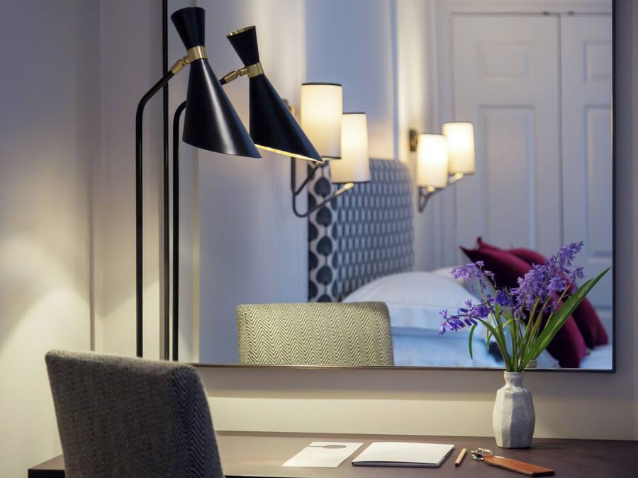 A hotel room with a desk, chair, and a large mirror reflecting a bed and a vase with flowers.