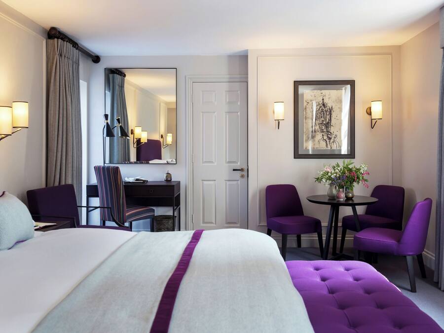 Ein Hotelzimmer mit einem großen Bett, einem Schreibtisch, zwei violetten Sessel, einem kleinen Tisch mit Blumen und einem gerahmten Bild an der Wand.