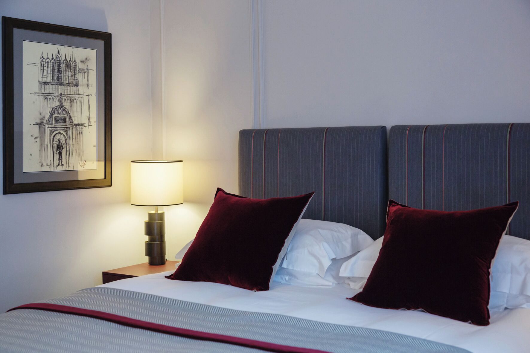 Hotellihuone, jossa on sänky, kaksi punaista tyynyä, yöpöytä lampun kanssa ja seinällä oleva kehystetty kuva.