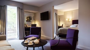 Habitación de hotel con cama, dos sillones morados, una mesita redonda con juego de té, un escritorio con silla, televisor de pantalla plana y un ventanal con cortinas.