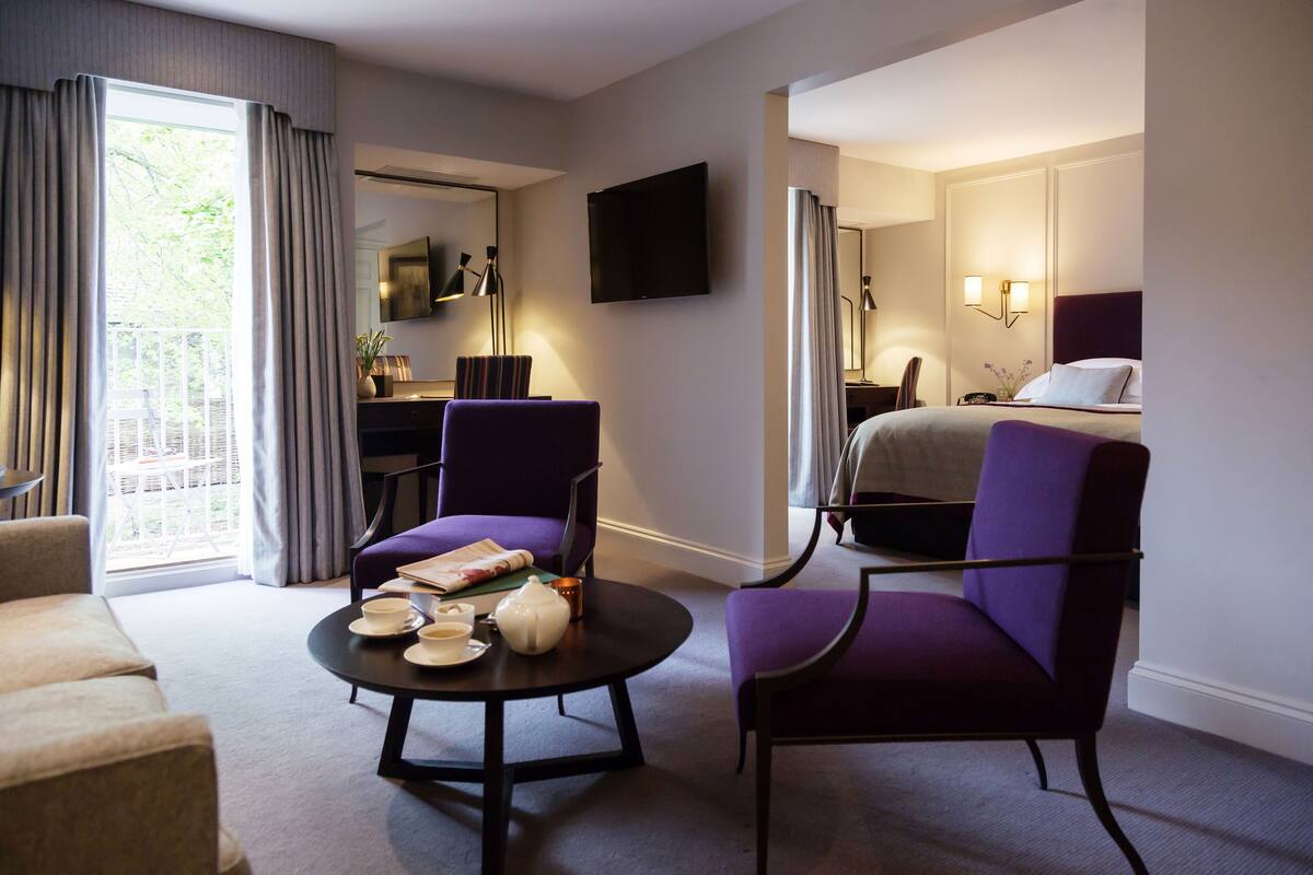 A hotel room with a bed, two purple armchairs, a round coffee table with tea set, a desk with a chair, a flat-screen TV, and a large window with curtains.