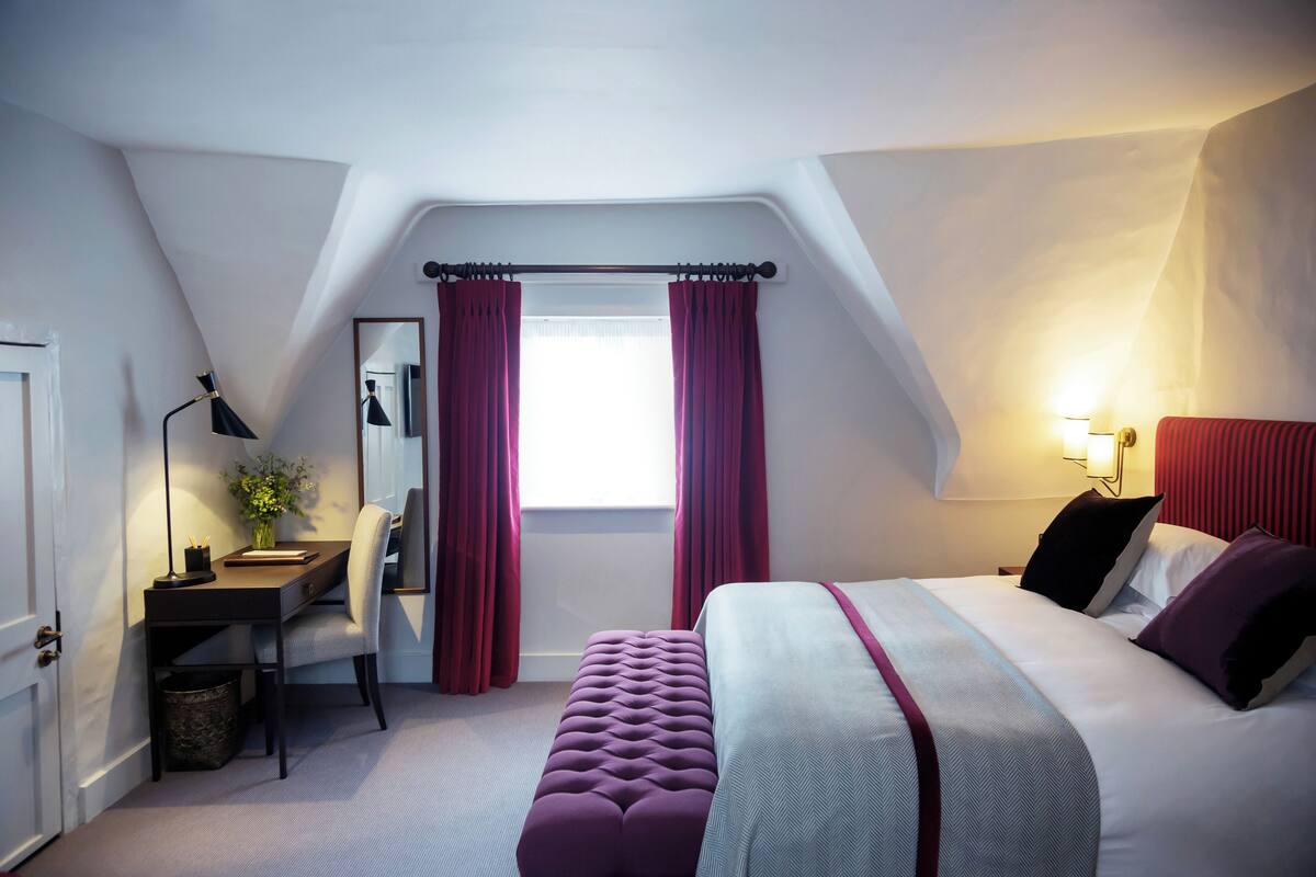 A bedroom with a bed, a desk, a chair, a window with curtains, and a lamp.