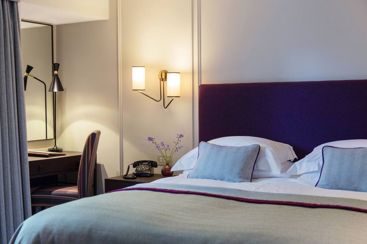 A hotel room with a bed, a desk with a lamp, a telephone, and a vase with flowers.