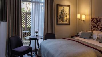 A hotel room with a bed, two chairs, a small table with a vase, a painting on the wall, and a view through a window with curtains.