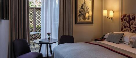 Habitación de hotel con cama, dos sillas, una mesita con un florero, un cuadro en la pared y vistas a través de una ventana con cortinas.