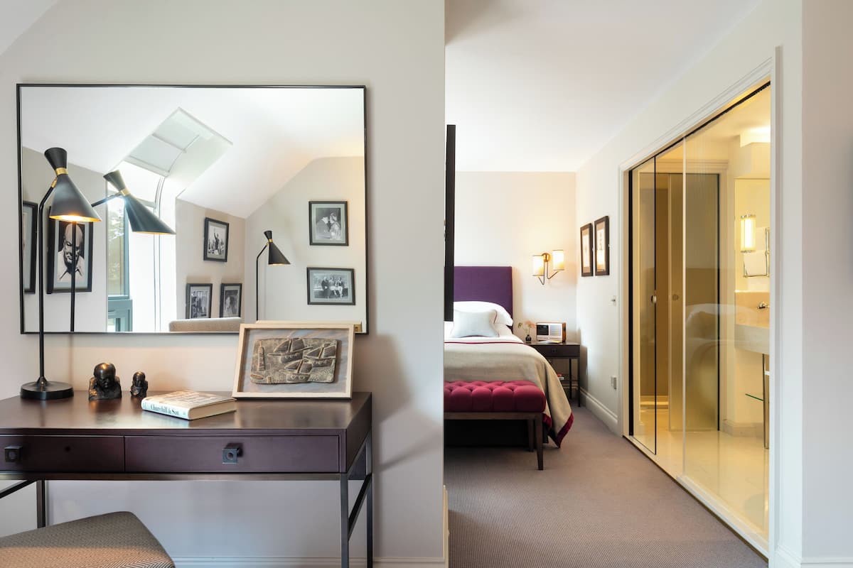 A modern hotel room with a large mirror, a desk with a lamp, a bed with a red accent, and a sliding glass door leading to a bathroom.