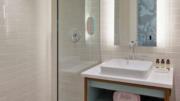 A modern bathroom with a glass shower enclosure, a white sink with a mirror above, and a wooden shelf with towels.