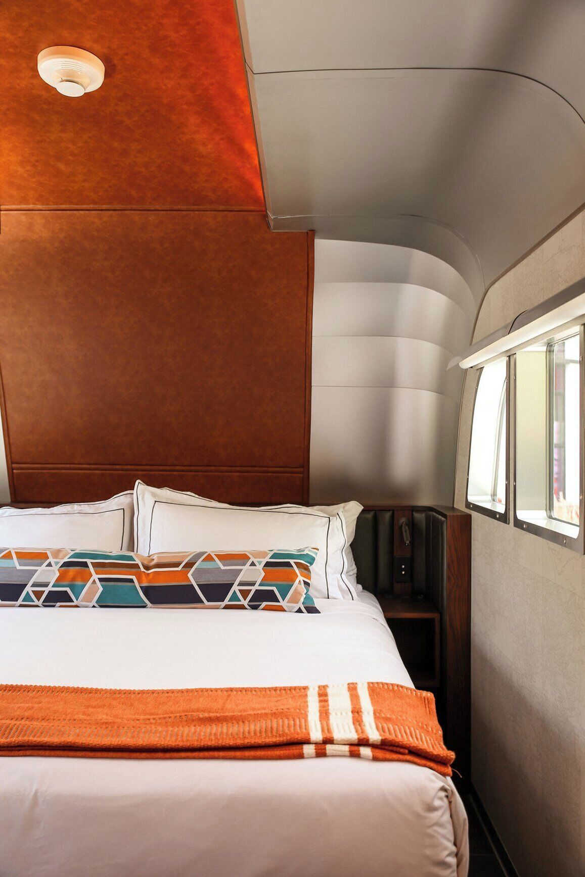 A bed with white linens, a colorful geometric pillow, and an orange throw blanket.