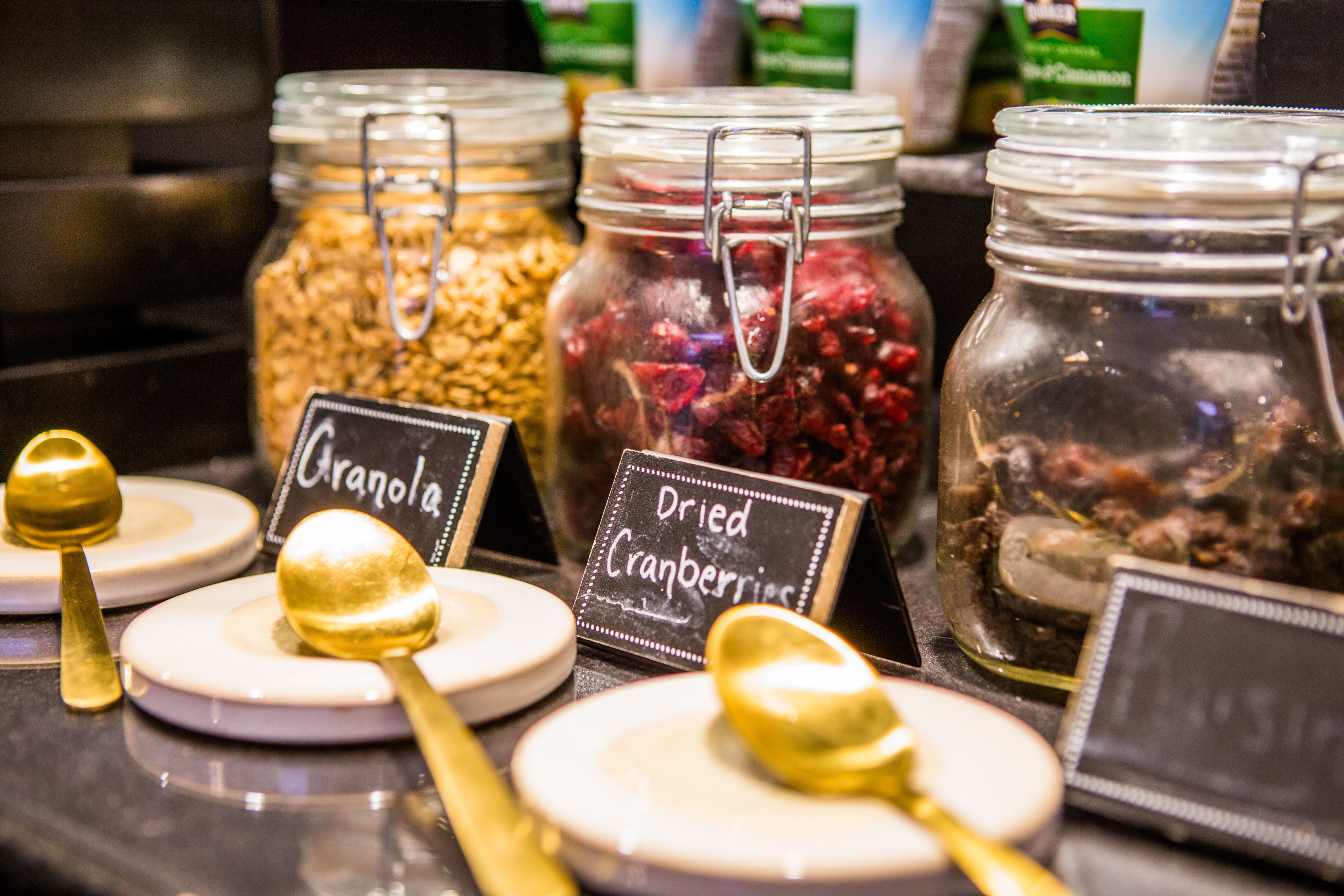 Une présentation de granola et de canneberges séchées dans des bocaux en verre, chacun muni d’une étiquette, le tout accompagné de cuillères sur des assiettes.