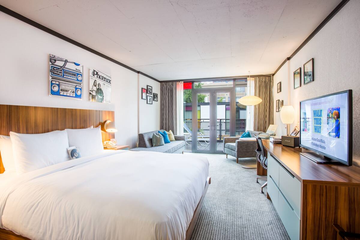 A hotel room with a large bed, a desk with a television, a sofa, and a balcony with outdoor seating.