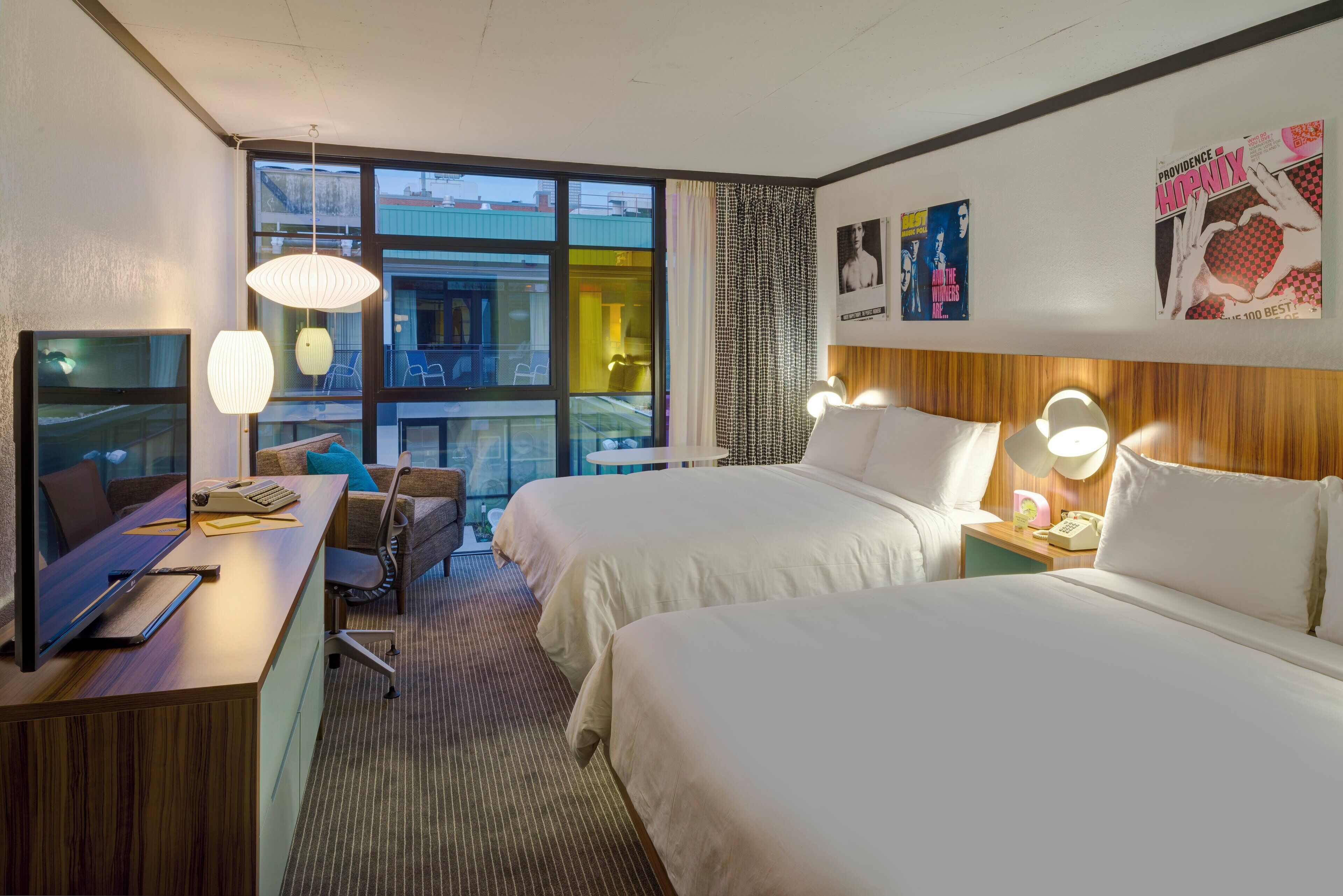 A hotel room with two beds, a desk with a computer, a chair, and a large window offering a view of the city.