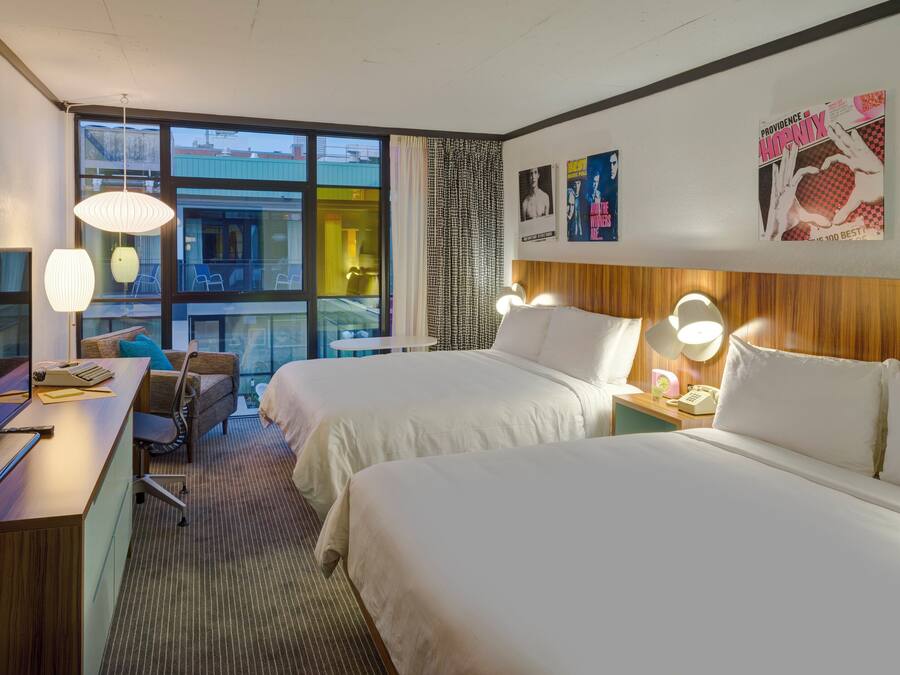 A hotel room with two beds, a desk with a computer, a chair, and a large window offering a view of the city.