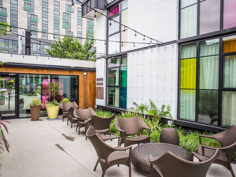 An outdoor seating area with wicker chairs and tables, surrounded by potted plants and a white building with large windows.