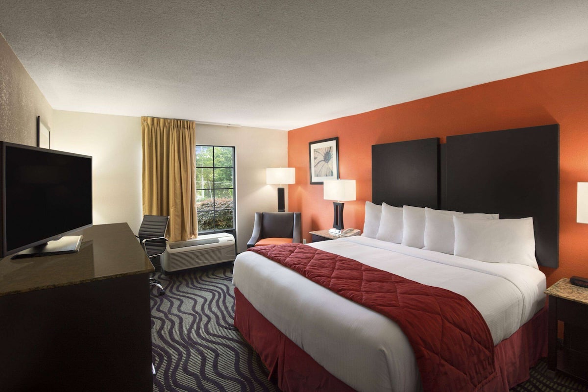 A hotel room with a large bed, a desk with a TV, a chair, a window with curtains, and a nightstand with lamps.