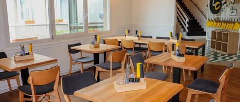 A restaurant with wooden tables and chairs, a staircase in the background, and a wall with yellow napkin holders.