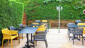 Un patio al aire libre con mesas y sillas, rodeado por una terraza de madera y un seto verde.