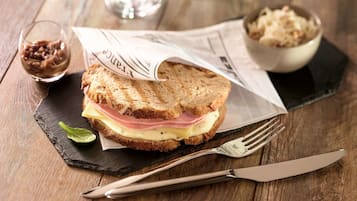 Un sándwich sobre una tabla de piedra negra, con un periódico debajo, acompañado de un tenedor, un cuchillo, un bol de chucrut y un vaso pequeño de salsa.