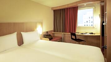 Habitación de hotel con una cama grande, un escritorio con silla, una ventana con cortinas y una mesita de noche con lámpara y bolso.