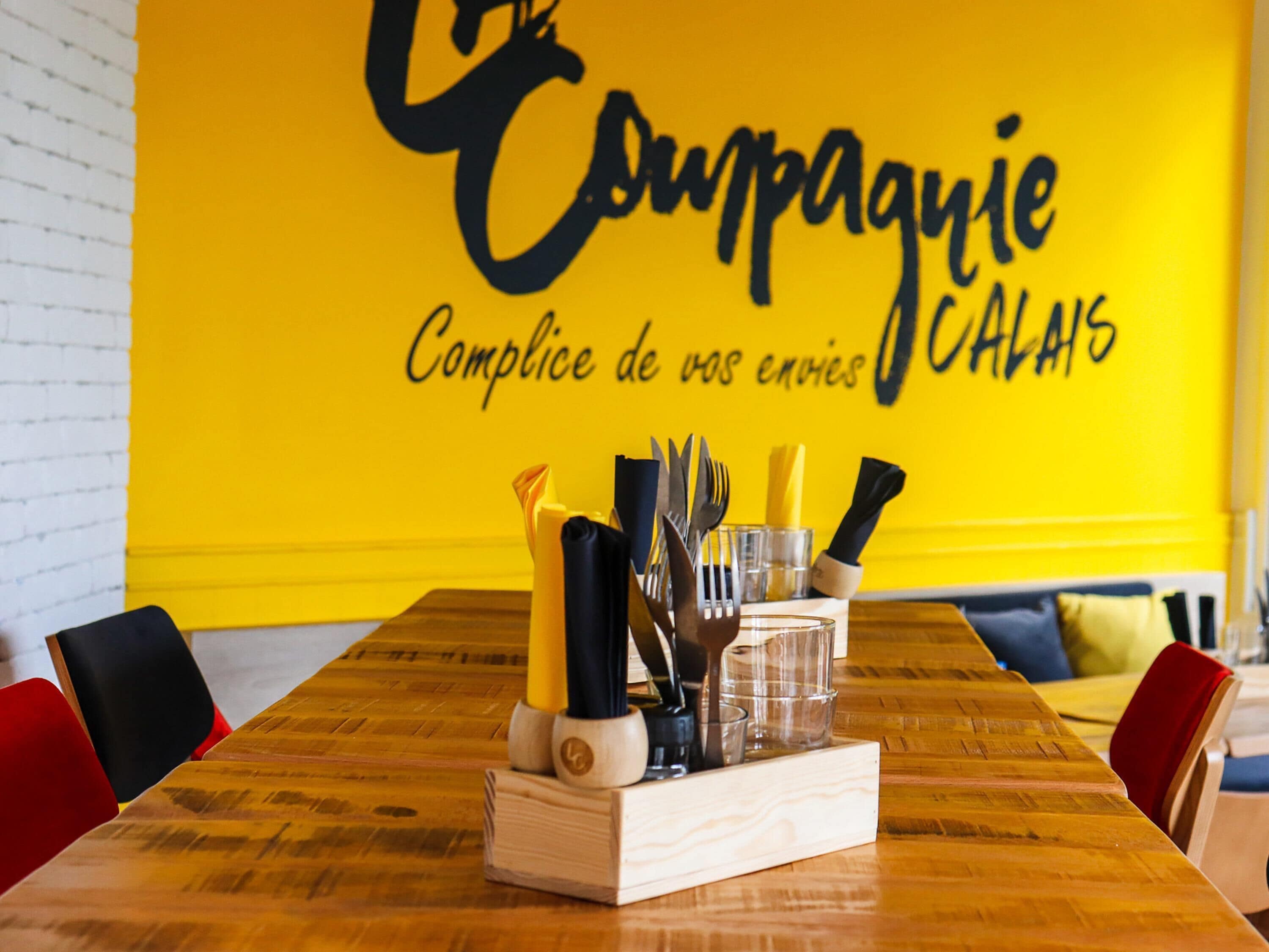 A dining table with cutlery and glasses, a yellow wall with text, and red and black chairs.