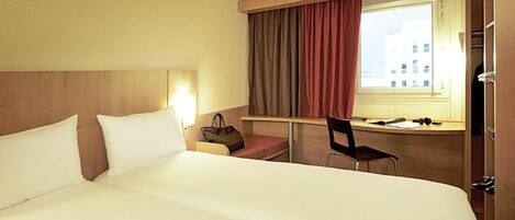 A hotel room with a large bed, a desk with a chair, a window with curtains, and a nightstand with a lamp.