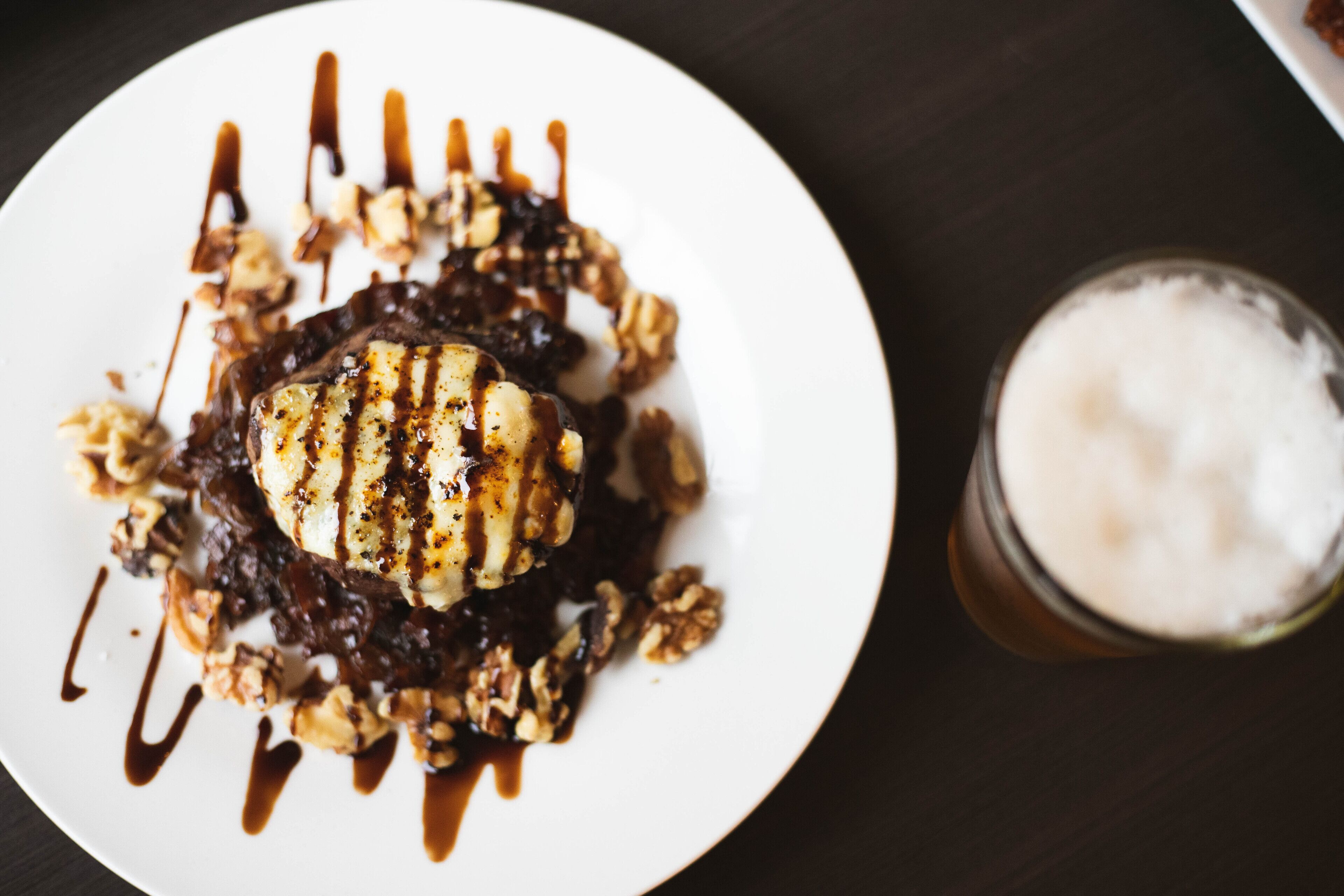 A dessert on a white plate with chocolate sauce and nuts, next to a glass of a creamy beverage.