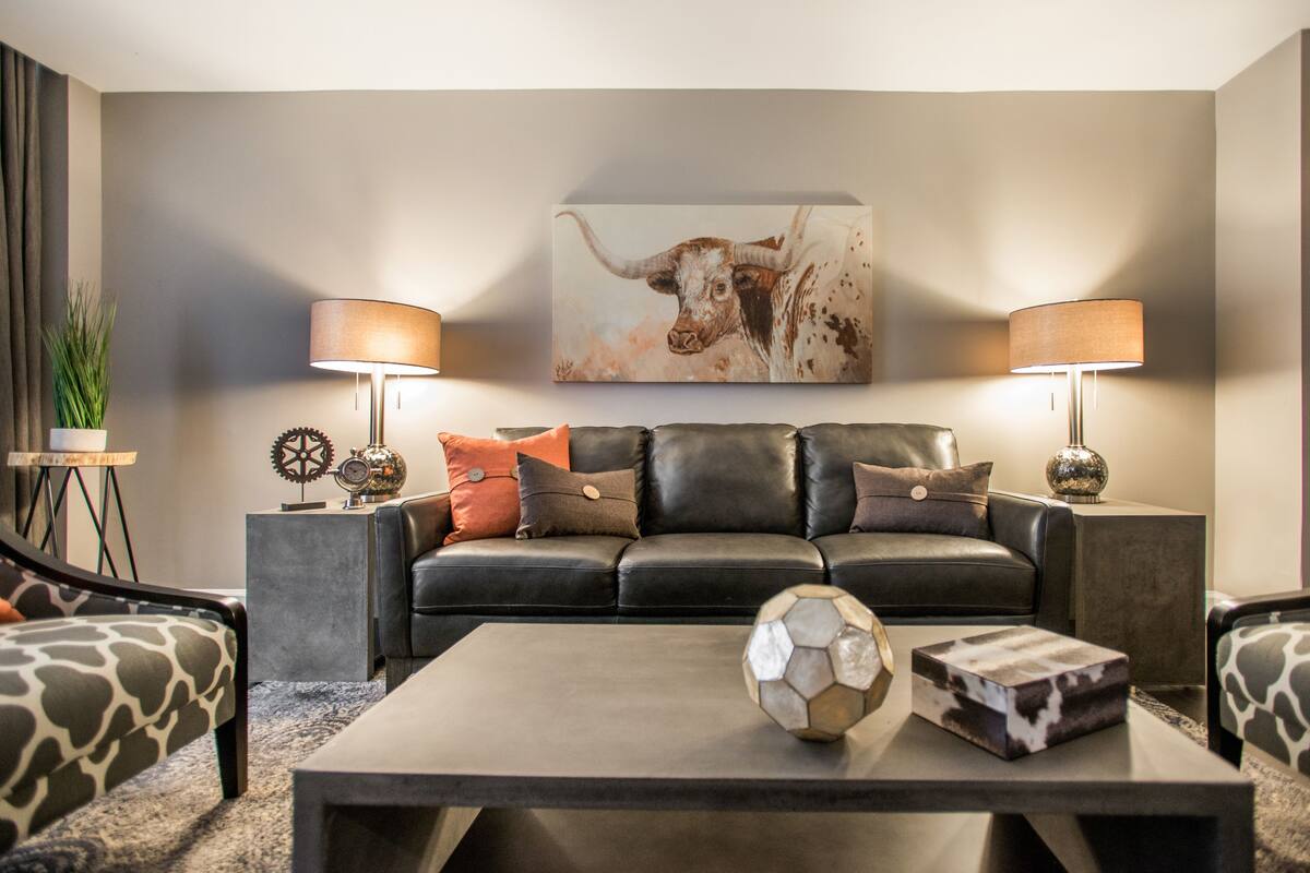 A modern living room with a black leather sofa, two side tables with lamps, a coffee table with a soccer ball and a decorative object, and a large painting on the wall.