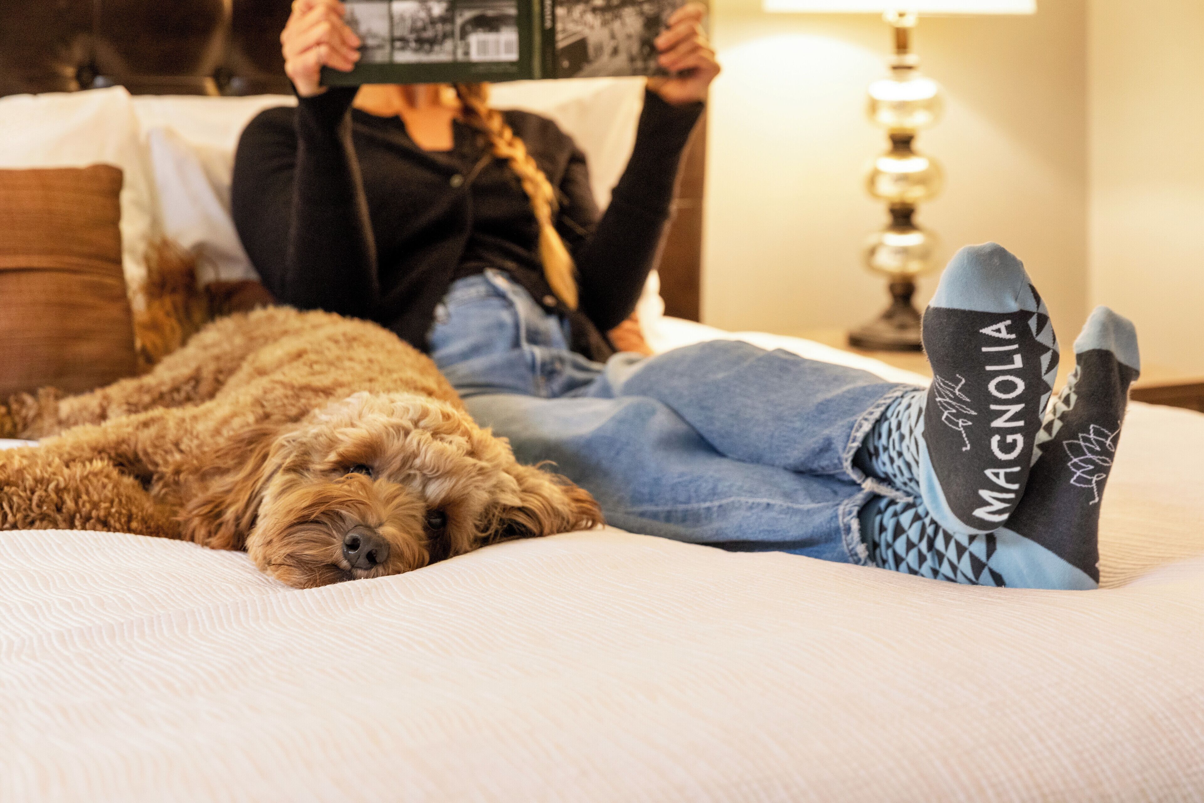 A person lying on a bed with a dog, holding a book, and wearing socks with a visible brand.