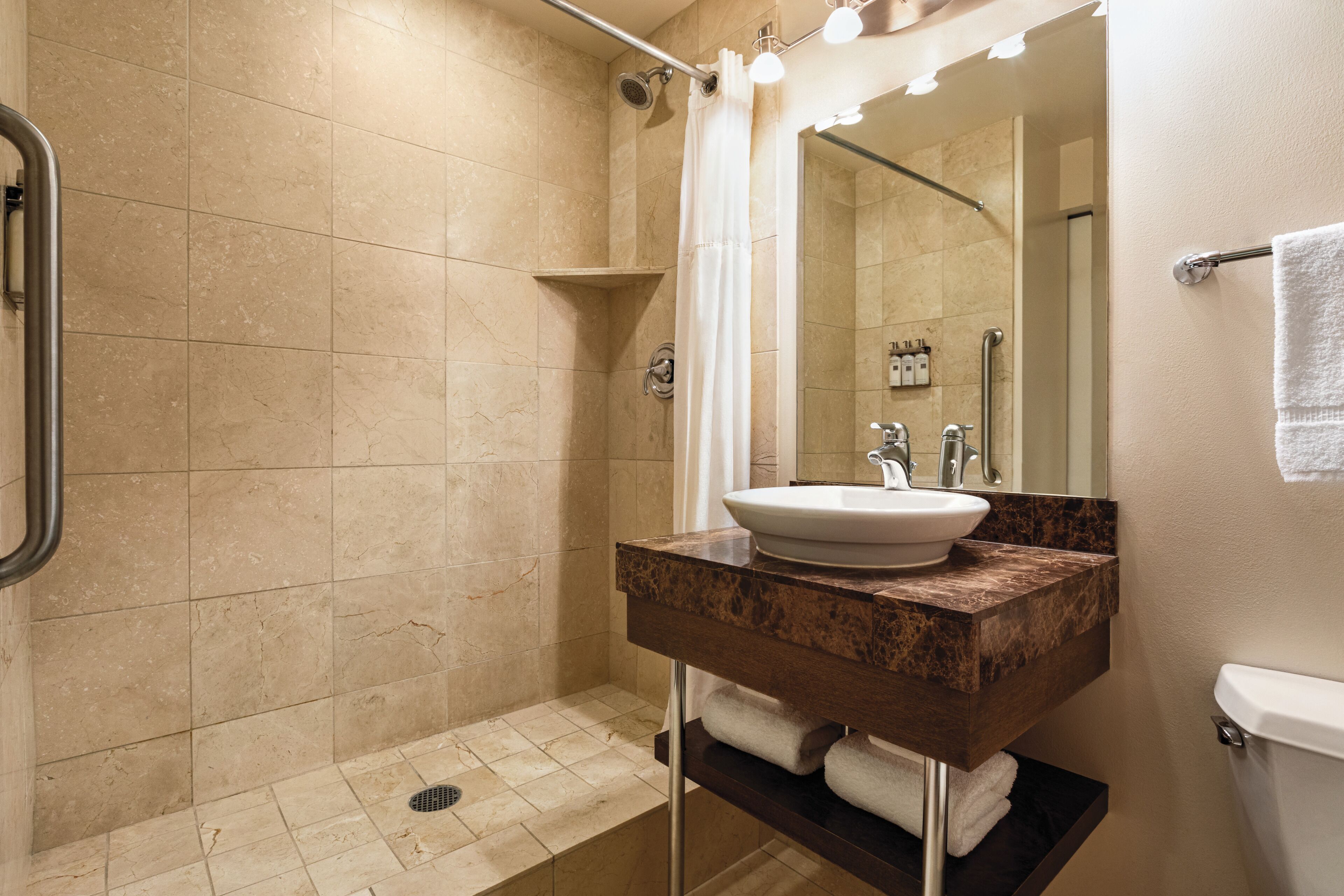 A modern bathroom with a beige tiled shower, a white sink with a marble countertop, and a mirror with lights above it.