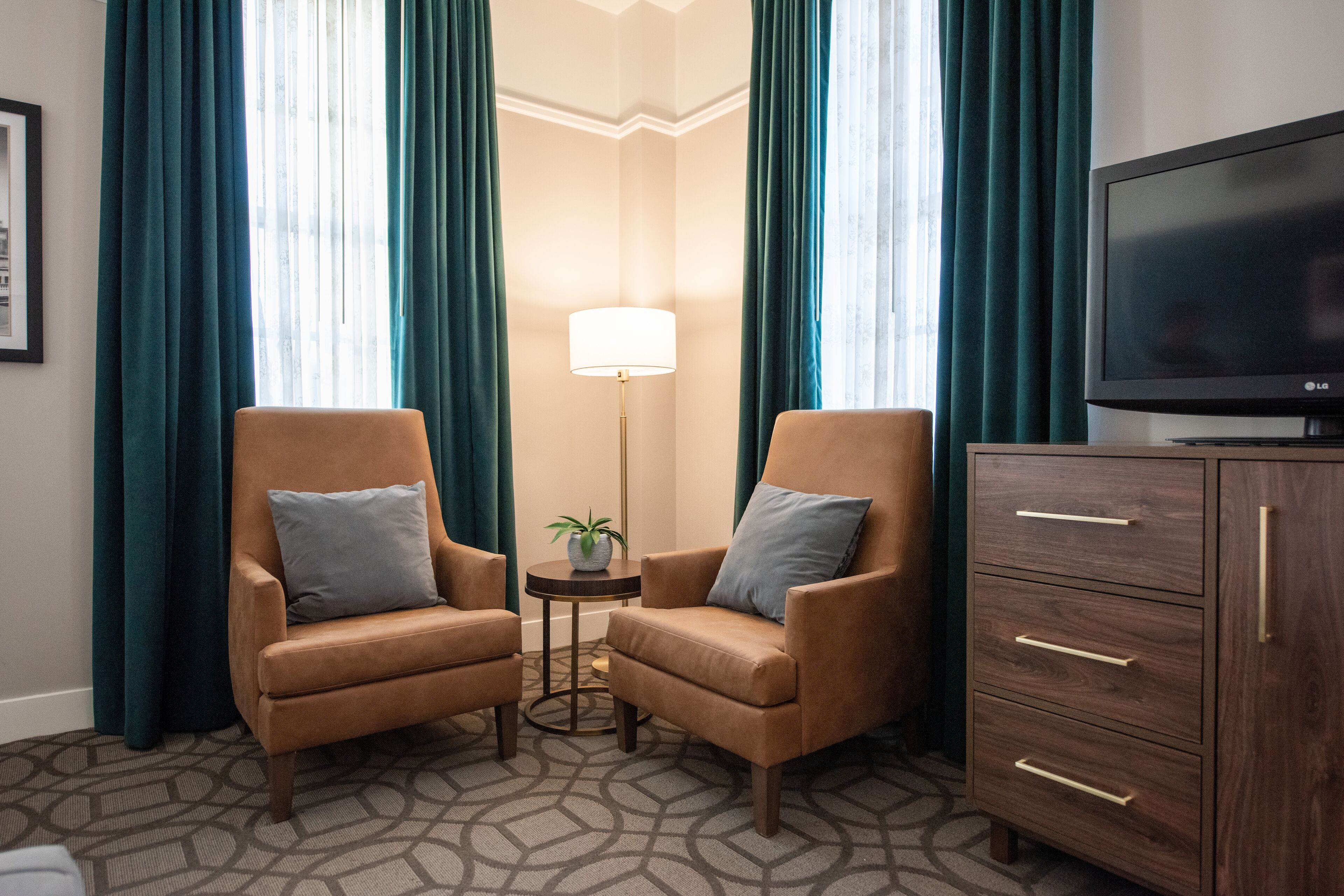 A hotel room with two brown armchairs, a small side table with a lamp, a television, and a wooden cabinet.