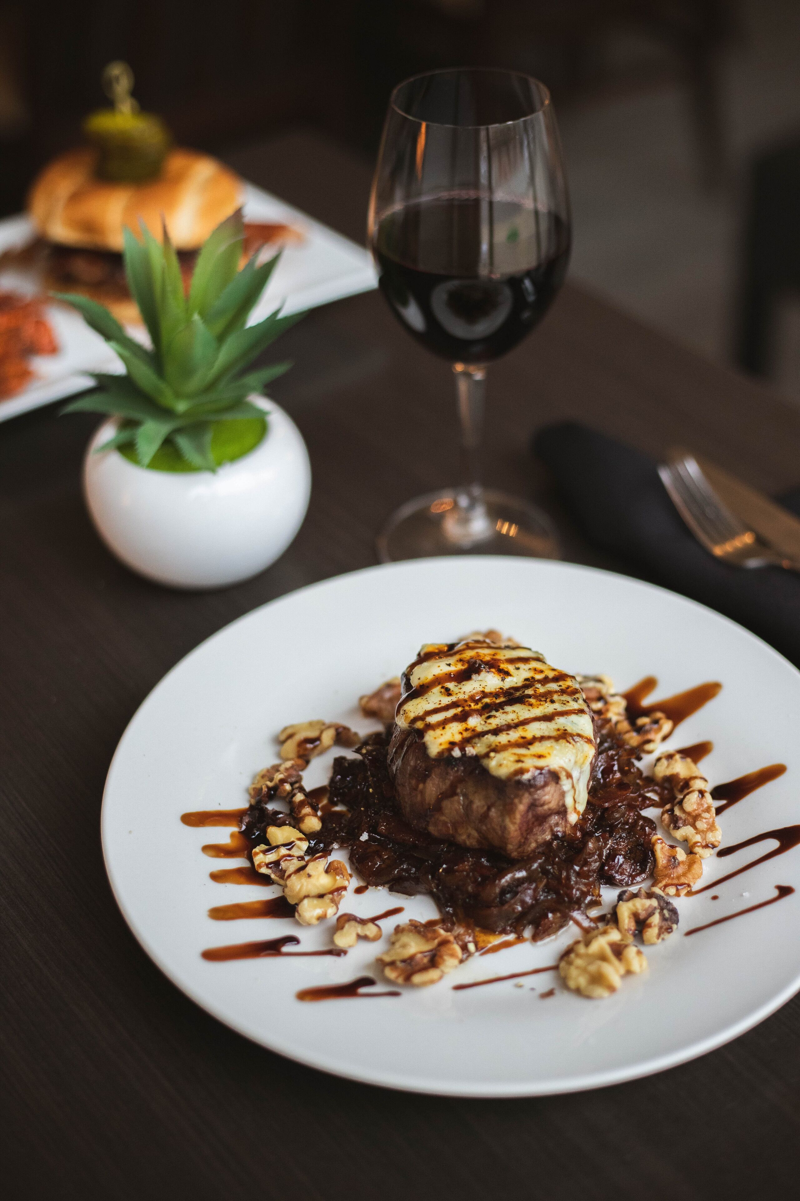 A plate of food with a piece of meat topped with sauce and walnuts, a glass of red wine, and a small plant in the background.