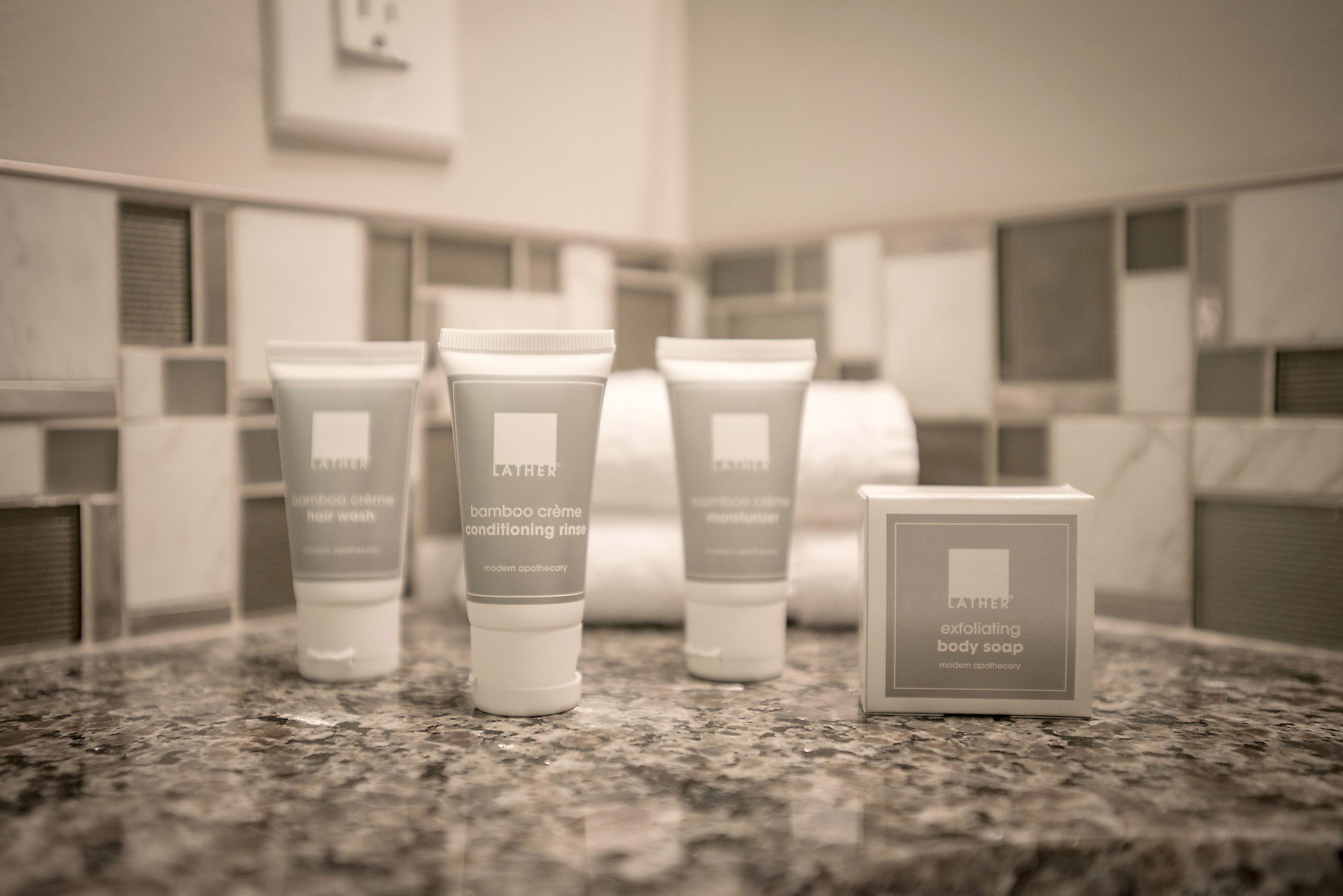 Une salle de bain dotée d’un comptoir en granit sur lequel sont disposés divers produits de toilette de la marque Lather, notamment un shampooing, un après-shampooing et un savon pour le corps.