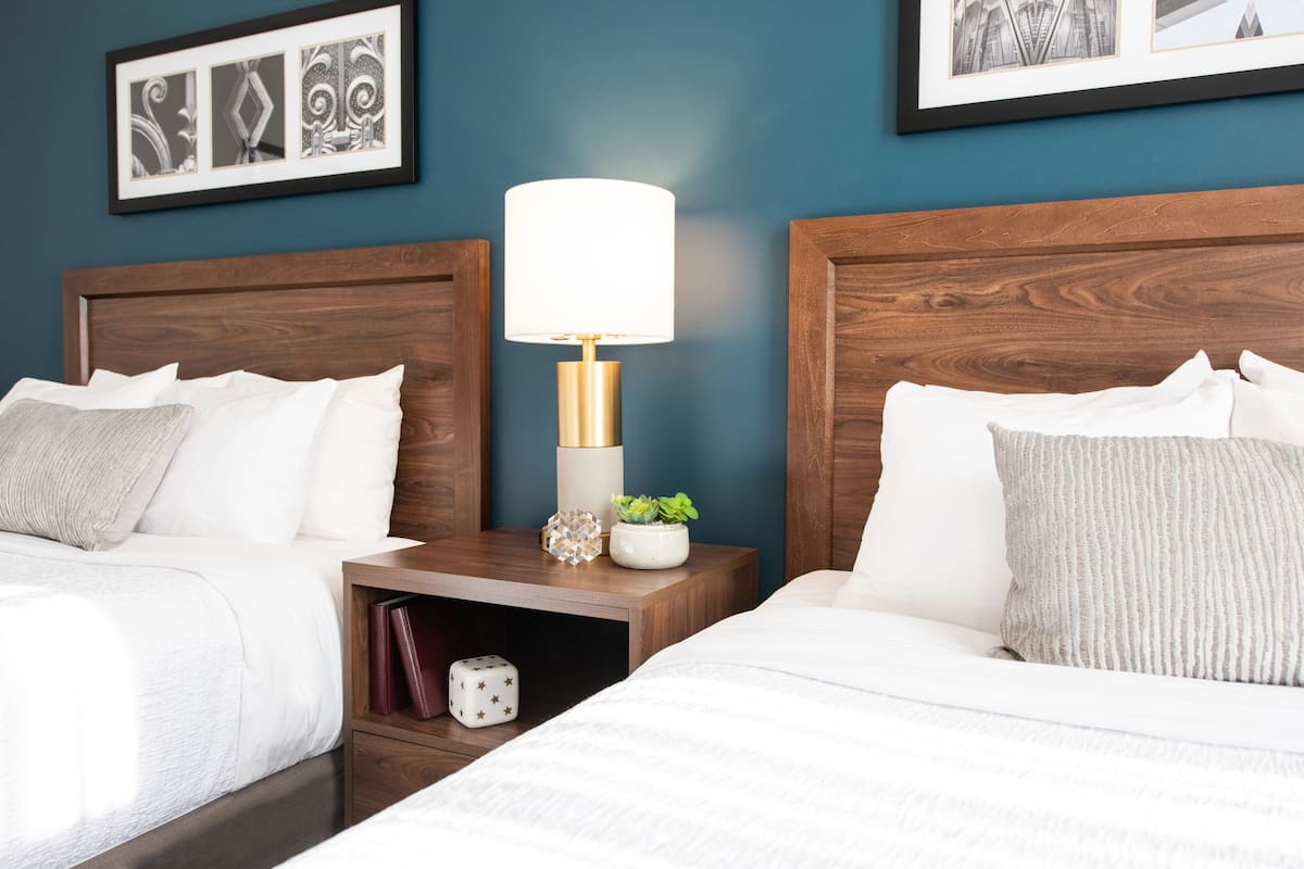 A hotel room with two beds, a nightstand with a lamp, framed pictures on the wall, and a small plant.