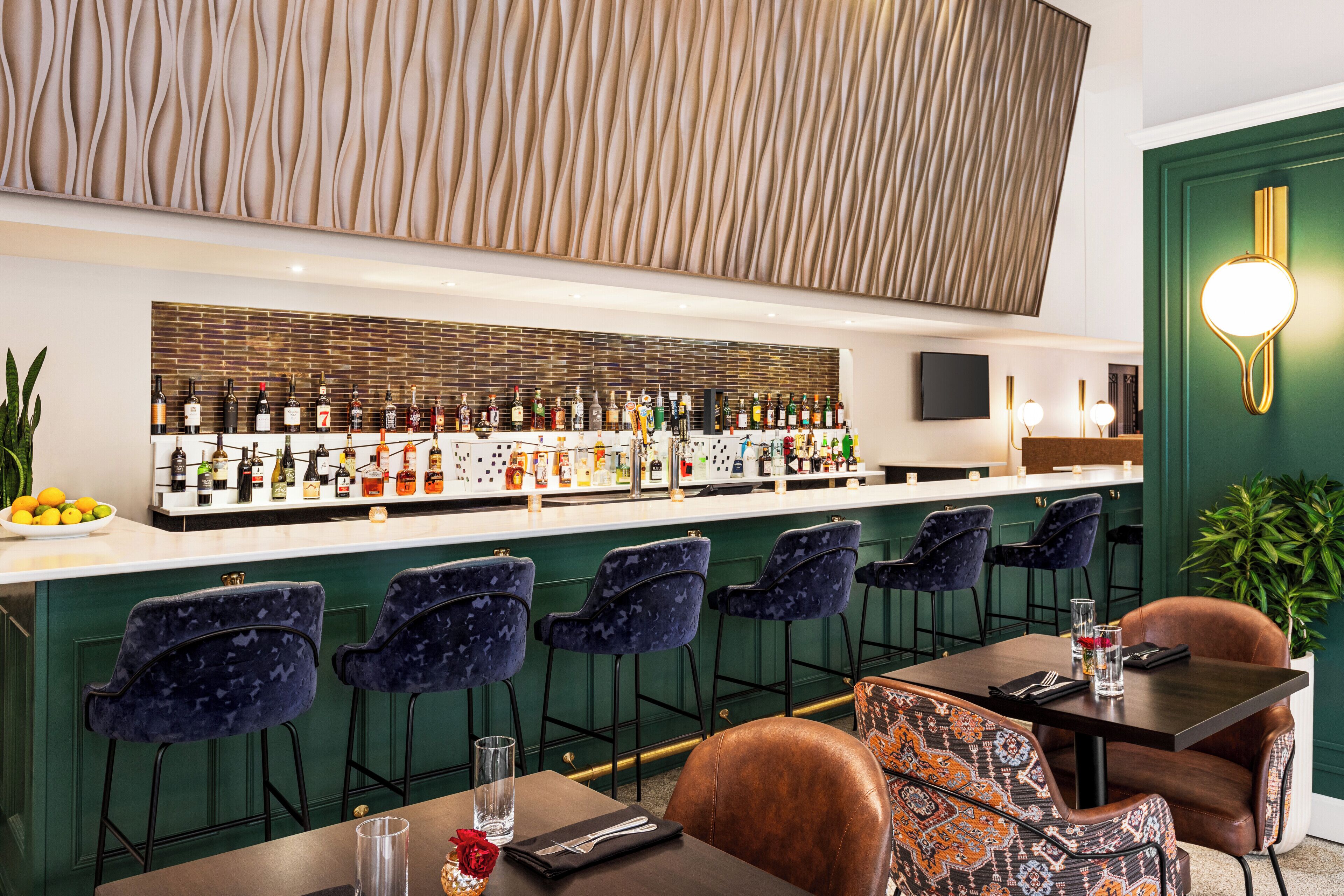 A bar area with a long counter displaying various bottles, blue upholstered bar stools, and a table with a brown leather chair.