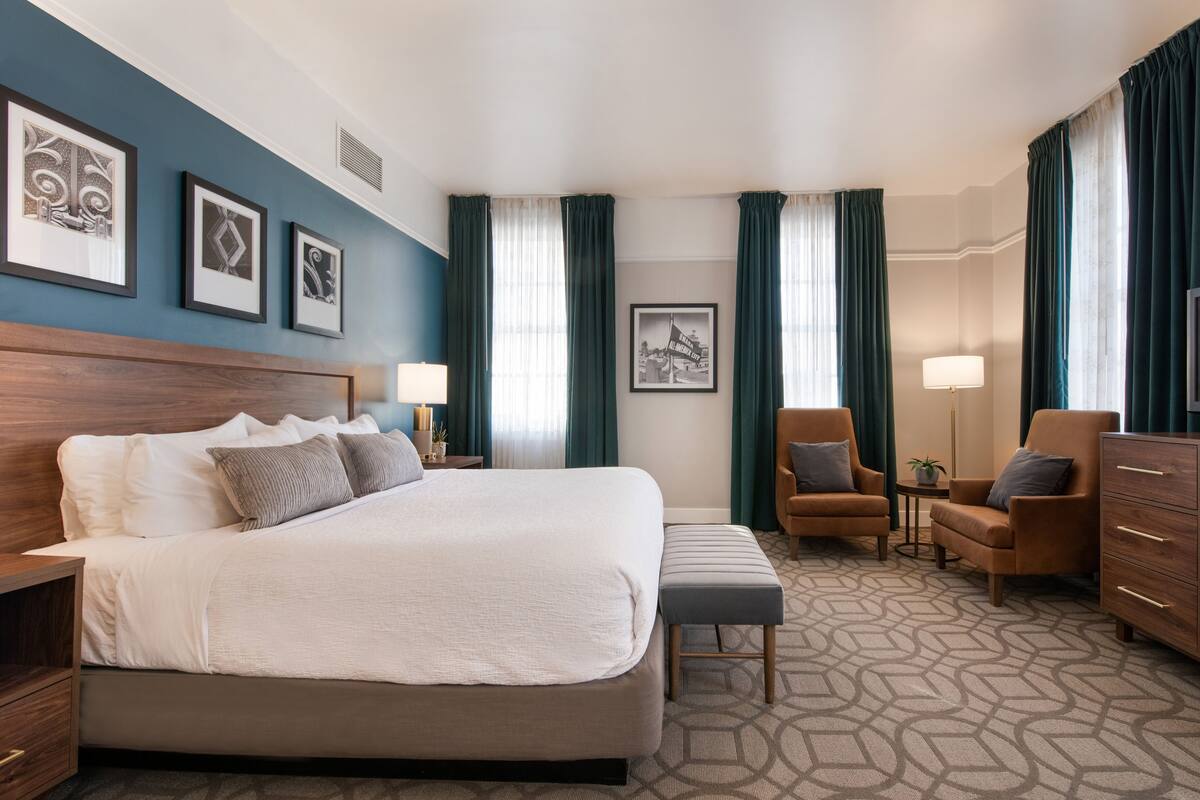 A hotel room with a large bed, two armchairs, a wooden dresser, and a small table with a lamp.