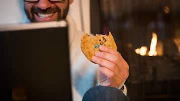 Une personne tenant un biscuit aux pépites de chocolat, assise devant un ordinateur portable.