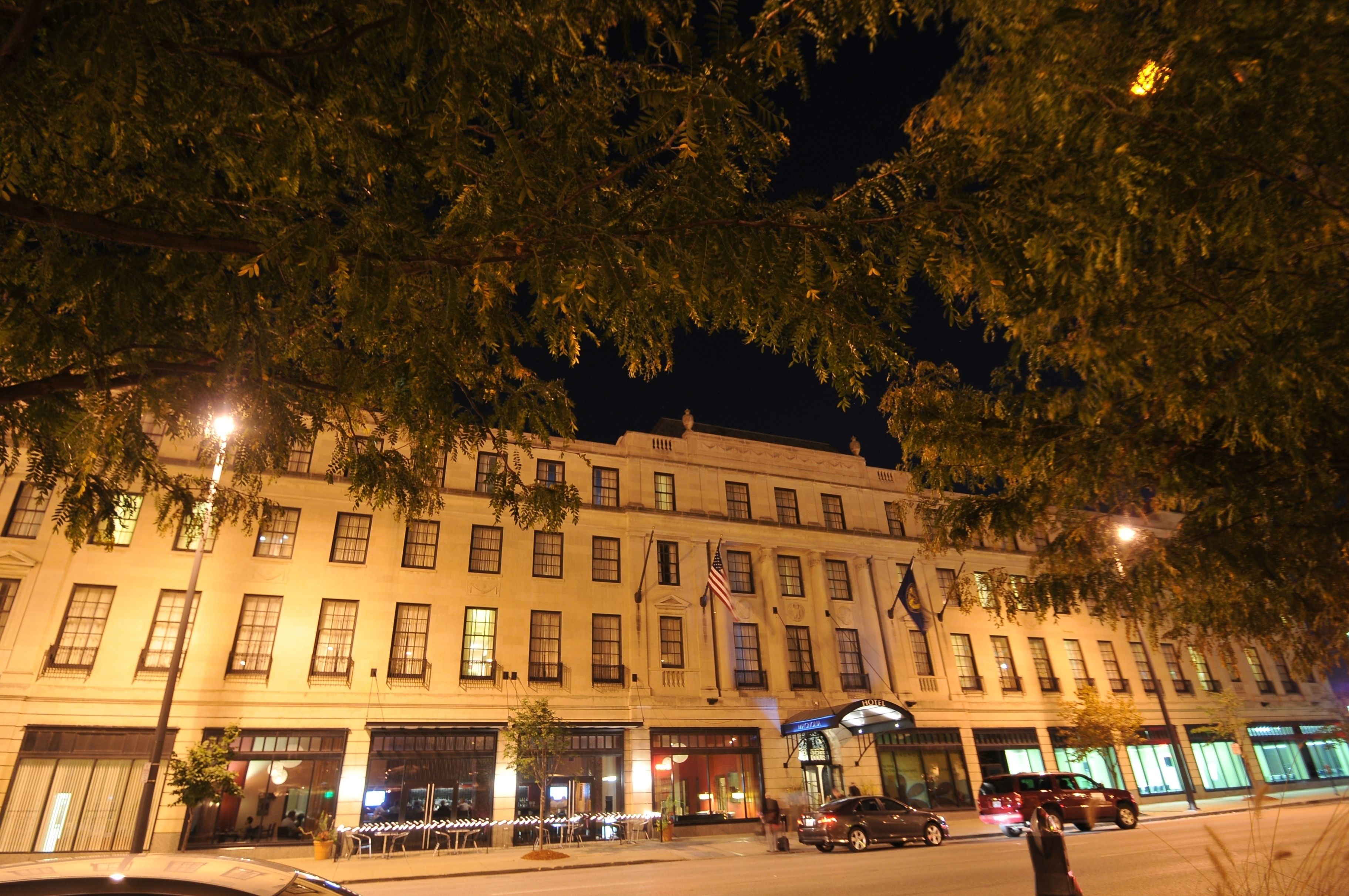 Un hotel de varios pisos con un estilo arquitectónico clásico, que presenta una entrada central con una bandera y una vista de la calle con coches aparcados y árboles.