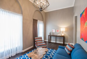 Un salon avec un canapé bleu, deux fauteuils, une table basse en bois, un lustre, une table d’appoint avec une lampe et un tableau coloré accroché au mur.