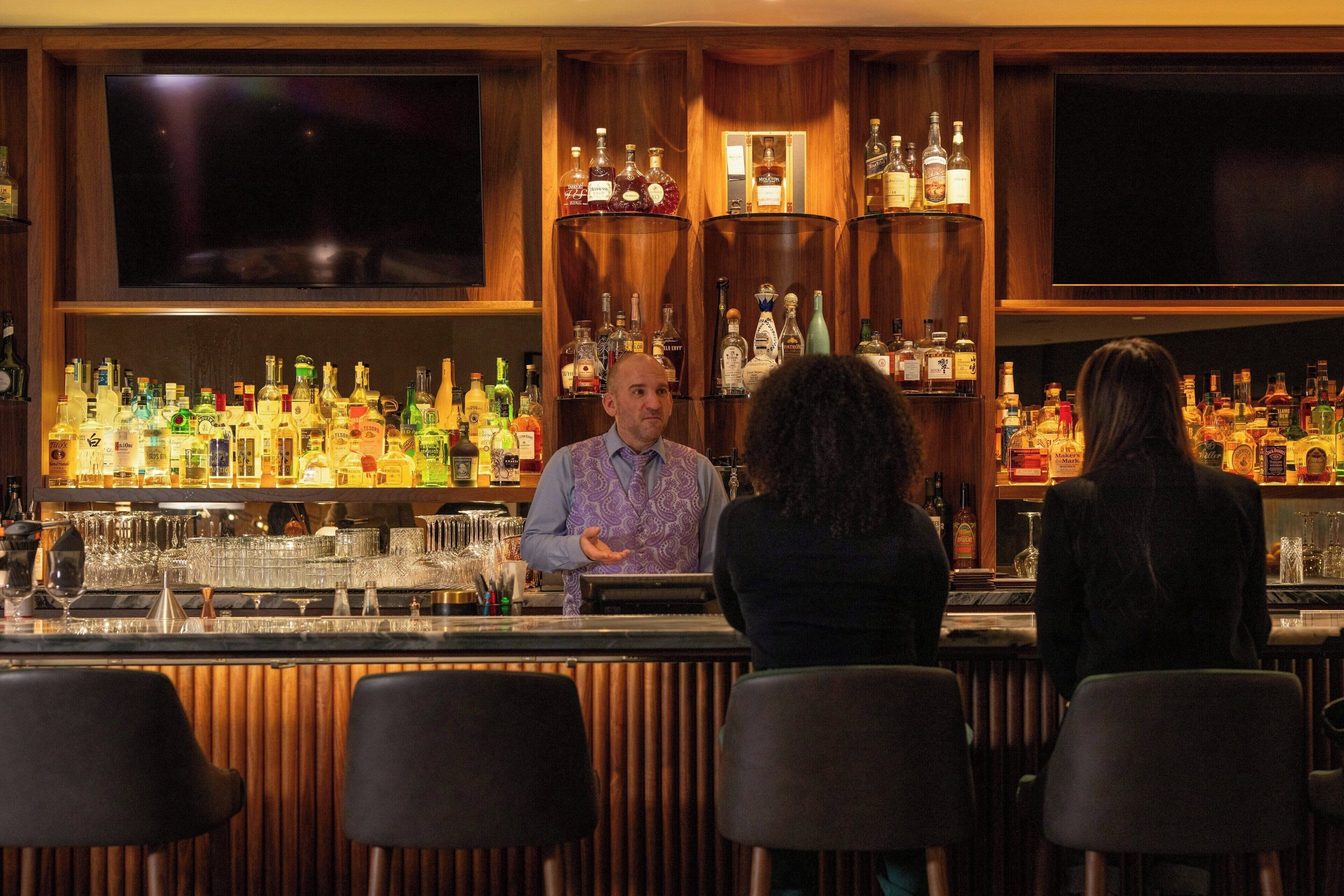A bar area with a bartender, shelves of liquor bottles, and bar stools.