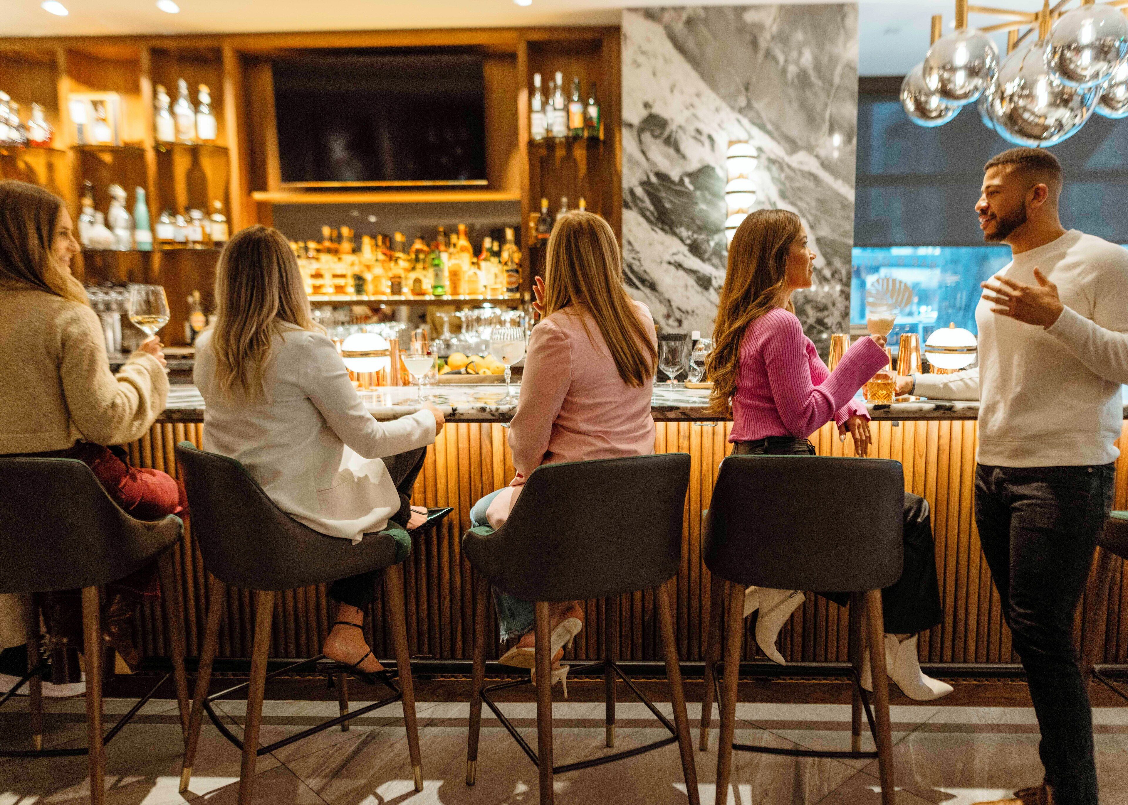 Eine Bar mit mehreren Tischen, an denen Gäste sitzen, einem Barkeeper, der sich um die Kunden kümmert, und einer Auswahl an Flaschen und Gläsern in Regalen.