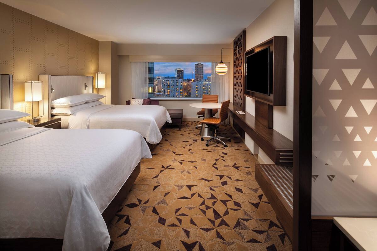 A hotel room with three beds, a dining table, a TV, and a city view.