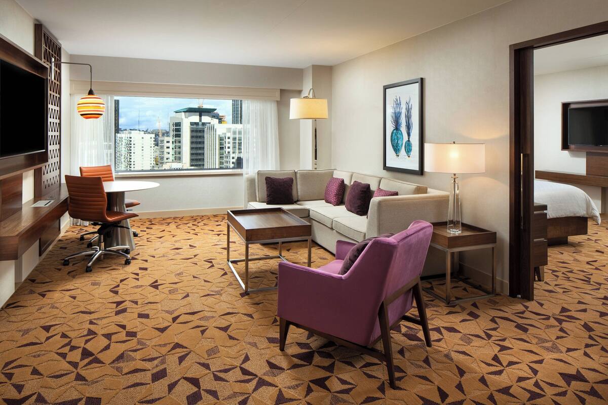 A hotel room with a sofa, armchair, and a small table. There is a TV, a lamp, and a window with a view of buildings.