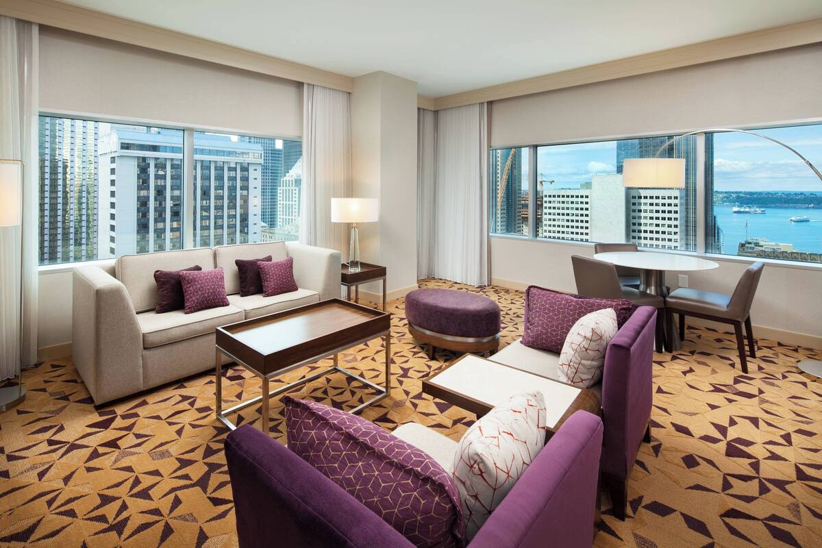 A modern hotel room with a sofa, ottoman, and armchair, a small table, and a view of the city and water.