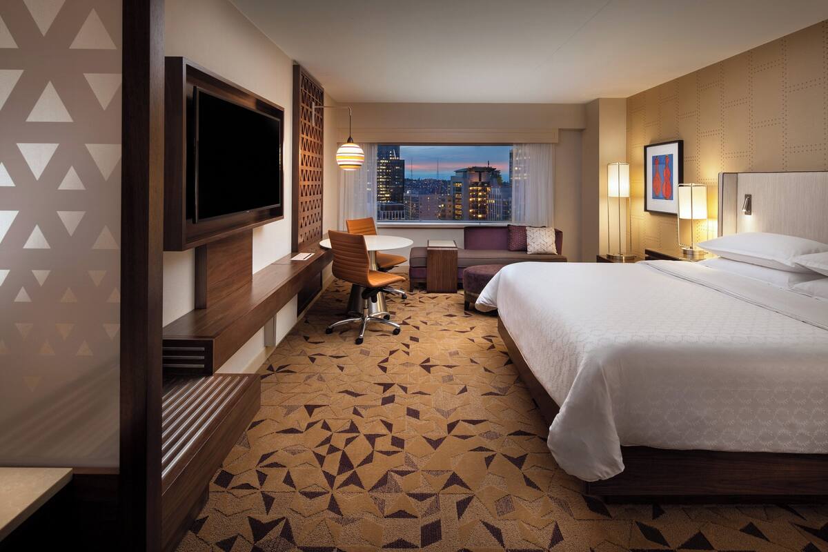 A hotel room with a large bed, a desk with a chair, a TV mounted on the wall, and a city view through the window.