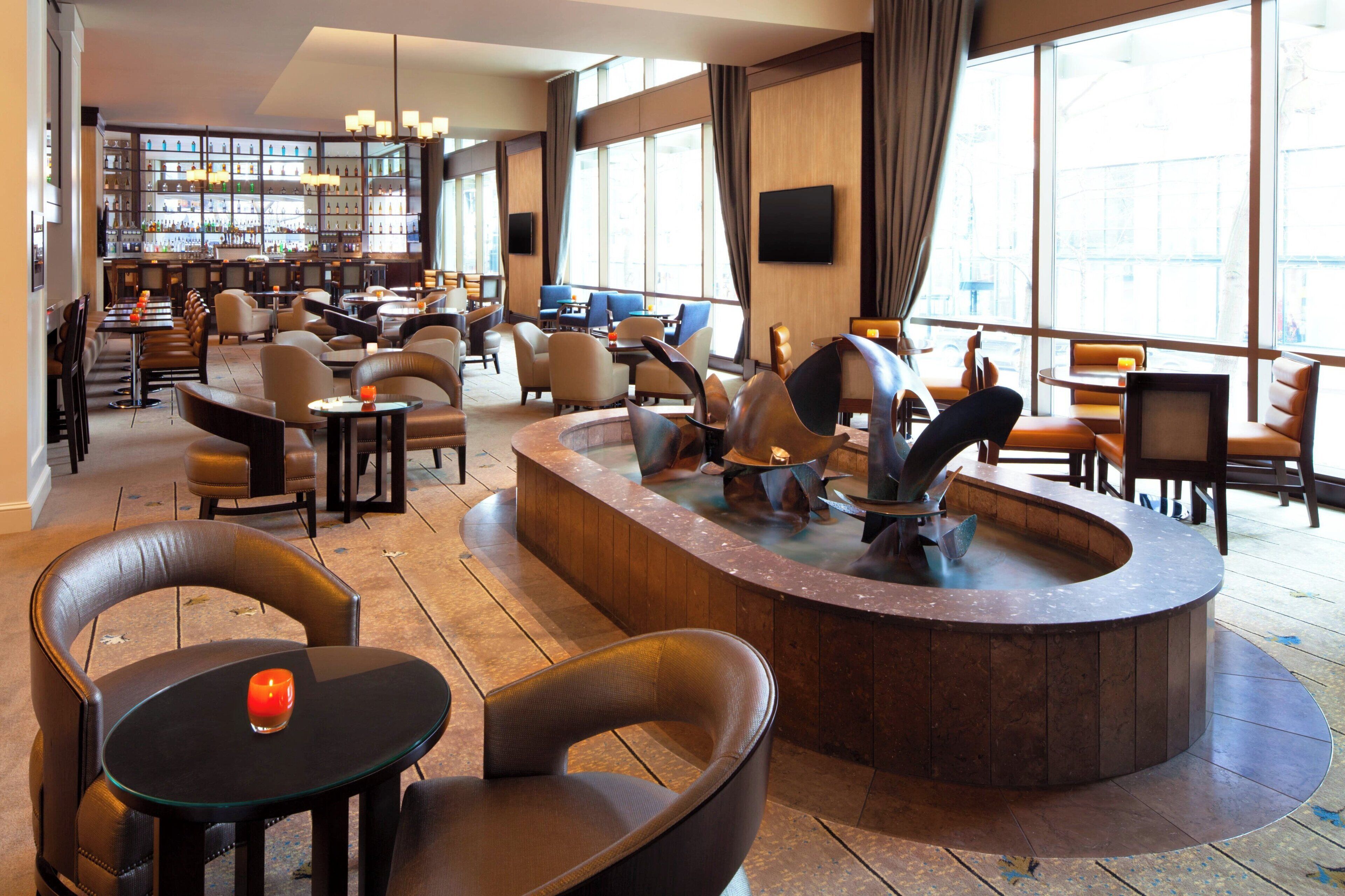 A spacious hotel lobby with multiple seating areas, a bar, and a large window offering a city view.