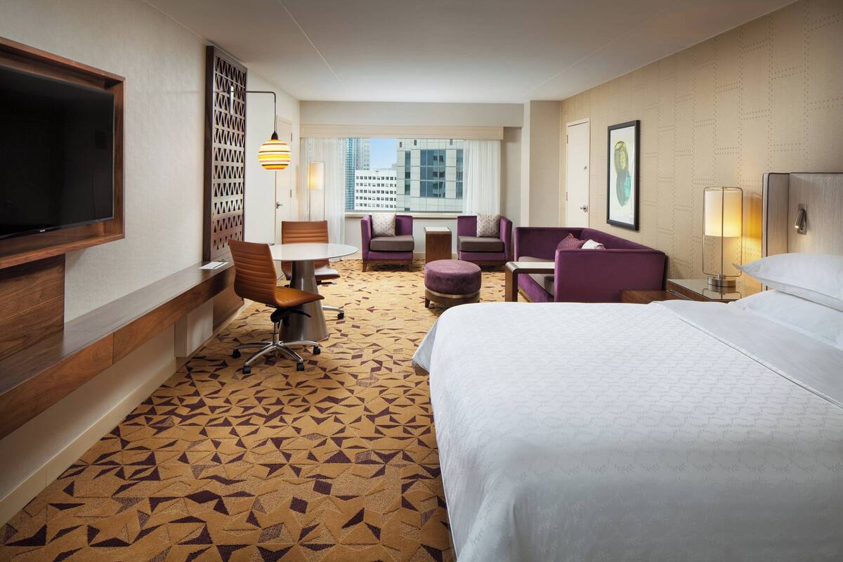 A hotel room with a large bed, a desk, a chair, and a sofa. There is a TV mounted on the wall, a window with a view of buildings, and a patterned carpet.