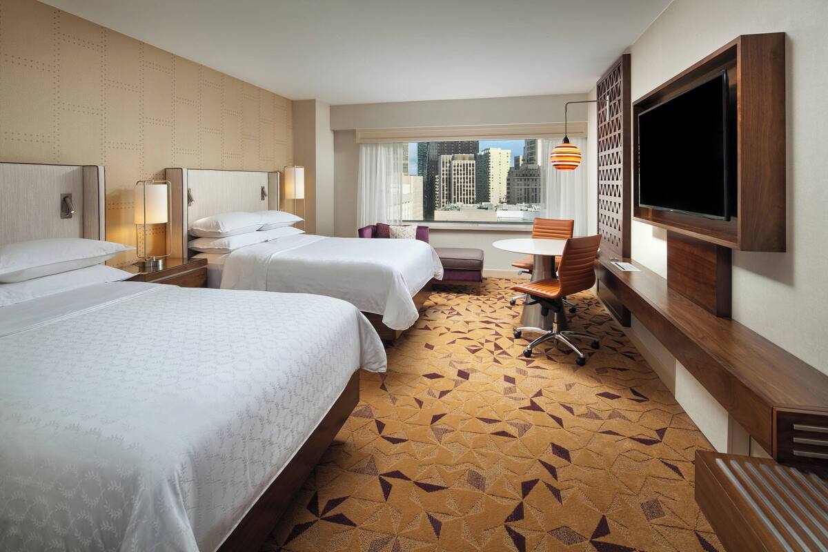 A hotel room with two beds, a desk, a chair, a TV, and a window with a city view.